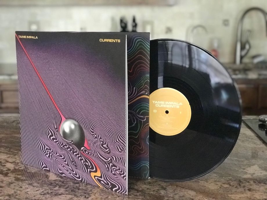 Tame Impala - Currents Vinyl LP with record protruding from the colorful, psychedelic artwork. The record is black with an orange-tinted middle that reads " Tame Impala/Currents"